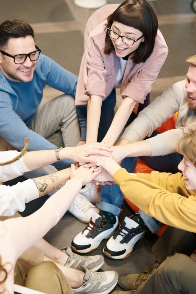 A diverse group of adults sit indoors, joyfully stacking hands together, symbolizing teamwork and camaraderie.